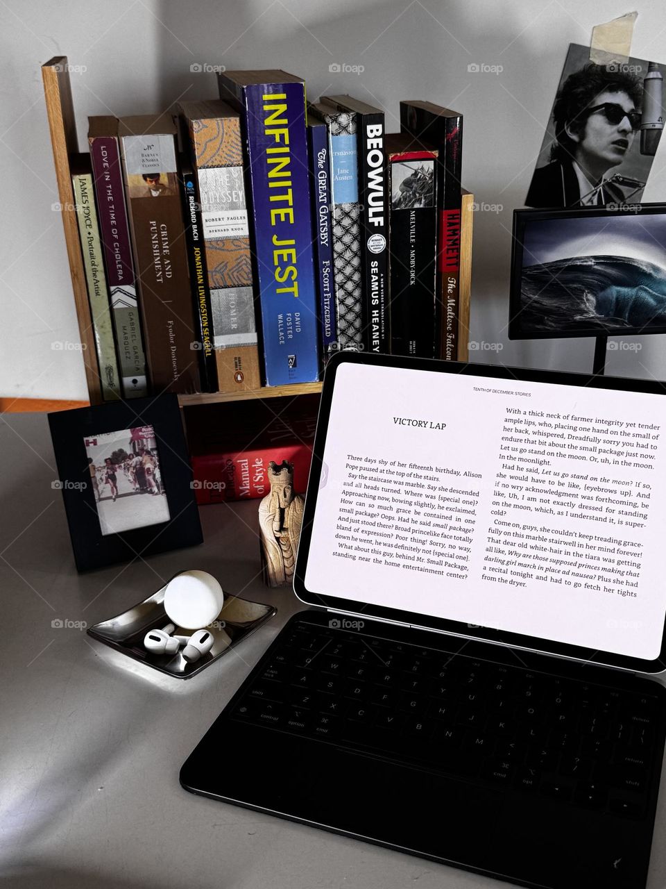 Desk with literary choice of books on bookshelf with iPad open to George Saunders story, with photos, ping pong ball, earbuds and reproduction Isle of Lewis chess queen