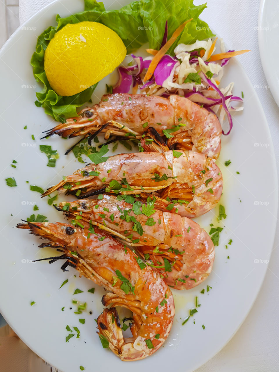Dish of grilled shrimps with vegetables salad, lemon and parsley on white plate at the table.