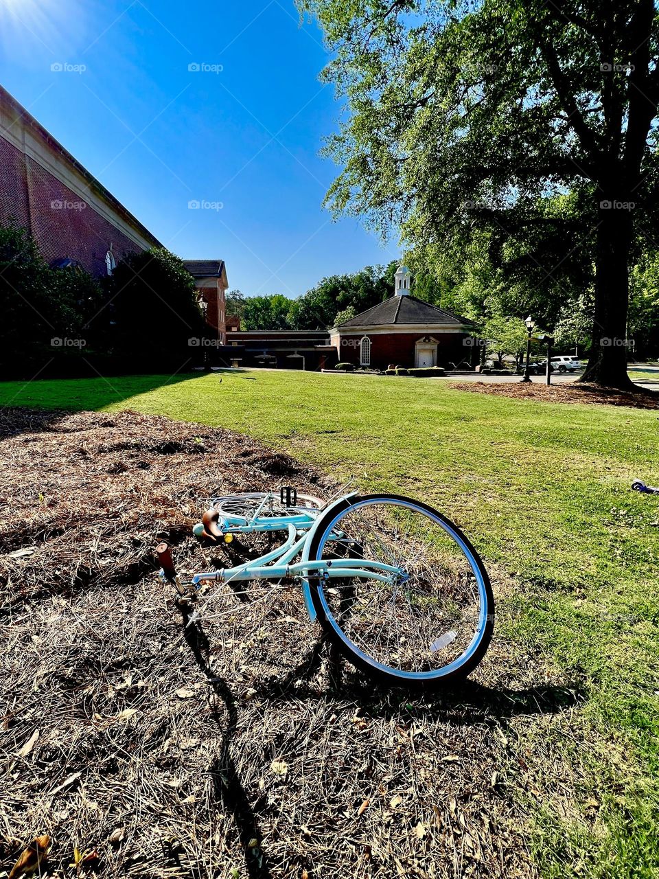 A blue cruiser style bicycle lying on its side in the foreground, with a grassy lawn, tree and brick church building in the background.