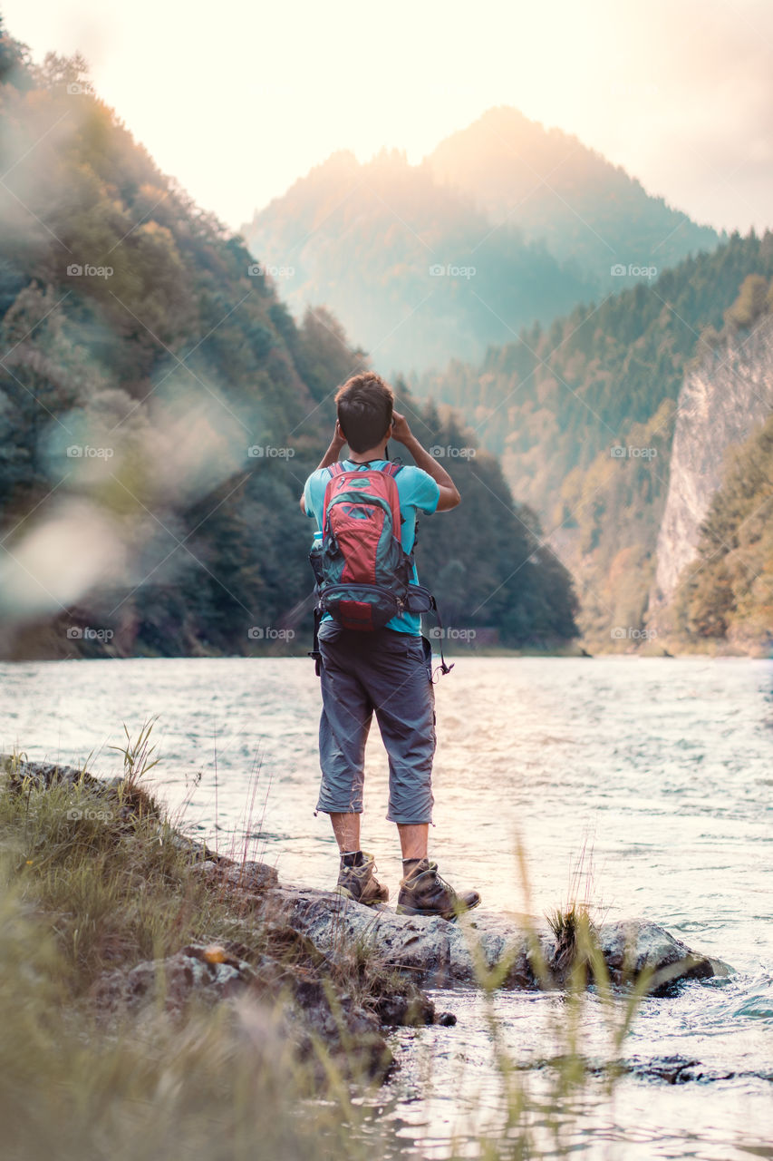 Young tourist with backpack looks through a binoculars on mountains peaks, stands on a rock over a river. Boy spends a vacation in mountains, wandering with backpack, he is wearing sports summer clothes