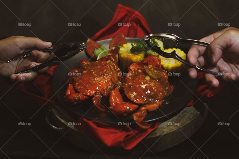 Crab flavored in creamy garlic sauce served with corn, tomatoes, lemon wedges and cherries on a black plate set on a red cloth set in a wooden setting. Asian flavor crab.