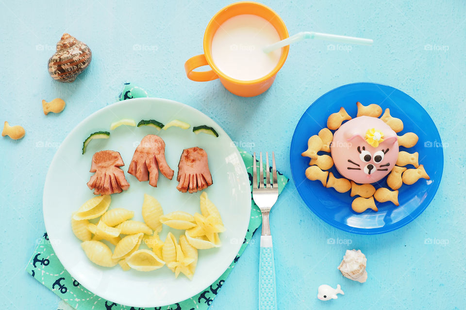 children's food serving. pasta in the form of shells and sausages in the form of jellyfish and octopus, cake cat and crackers fish, a Cup of milk.