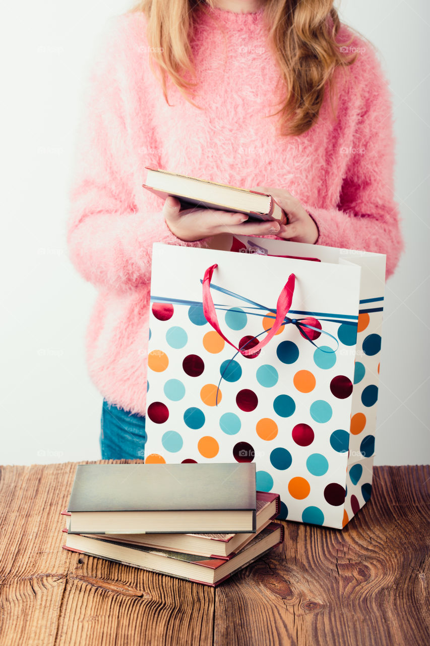 Young girl turning the pages of book in bookstore. Putting the books into paper bag. A few books on a wooden table. Teenager girl wearing pink sweater and blue jeans. Vertical photo