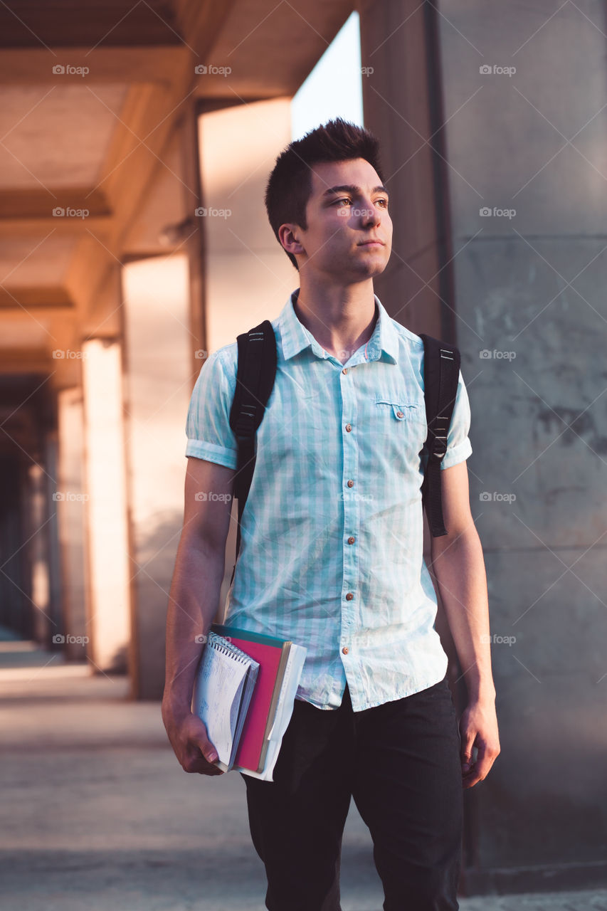 Student holding a notebook and carrying a backpack walking at the front of university building. Young boy wearing blue shirt and dark jeans