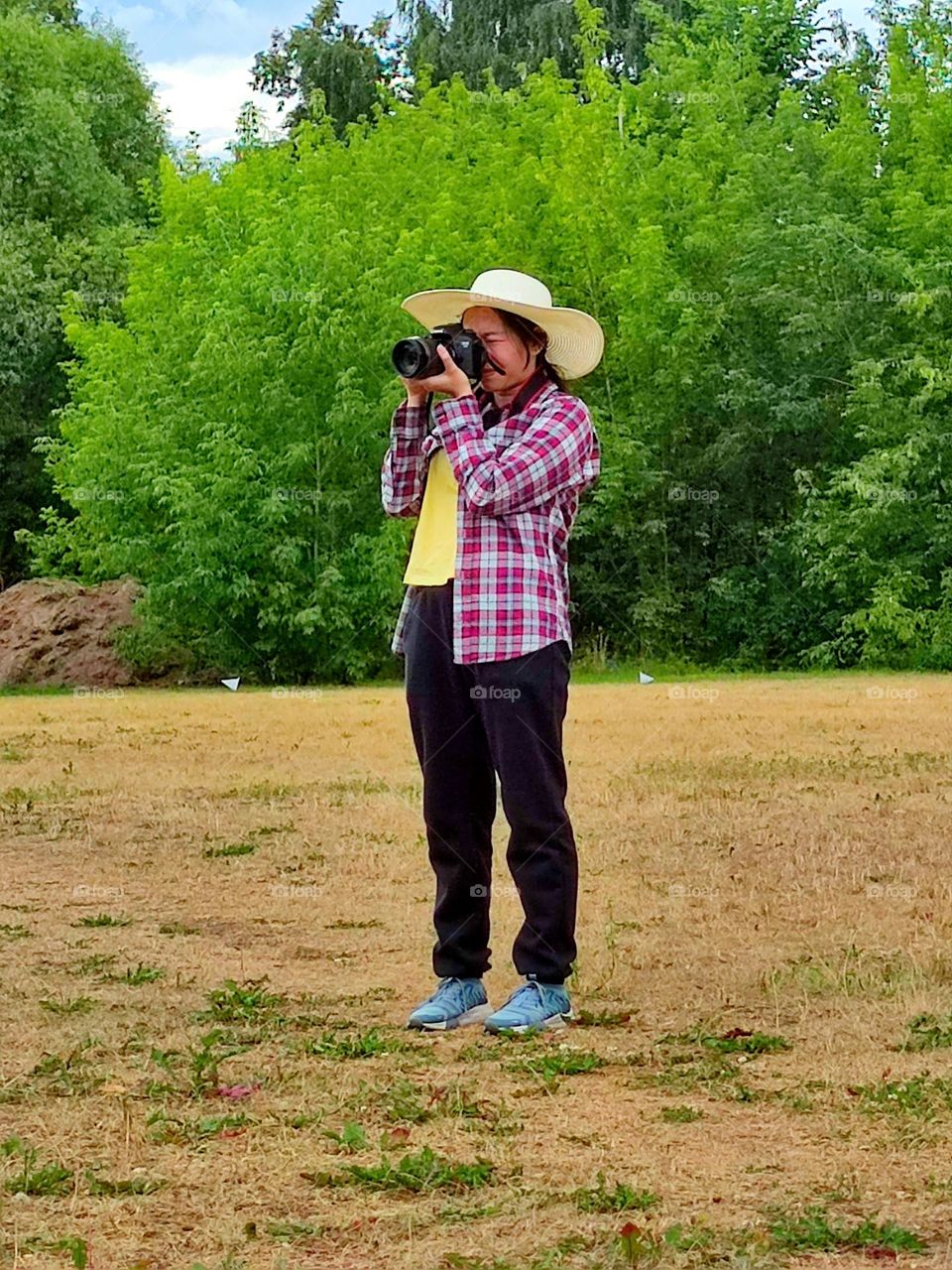 Summer day. A field surrounded by green trees. A girl in a straw hat stands on the field and takes pictures with a fotocamera