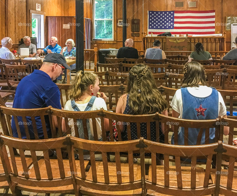 Small town life - enjoying a town BBQ and finding shelter inside the town hall from the rain. The American flag is prominent in the space.