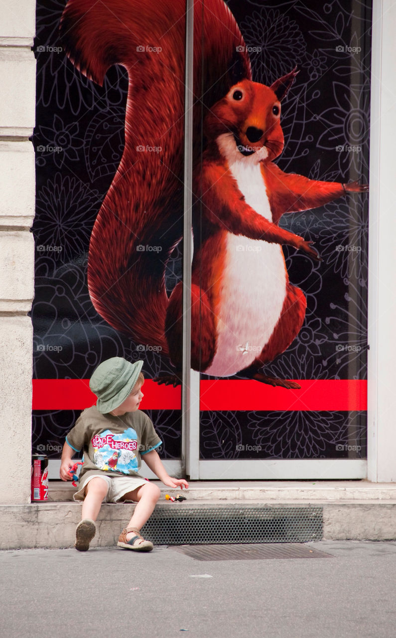 Small boy sitting on the curb in front of a poster of a giant squirrel