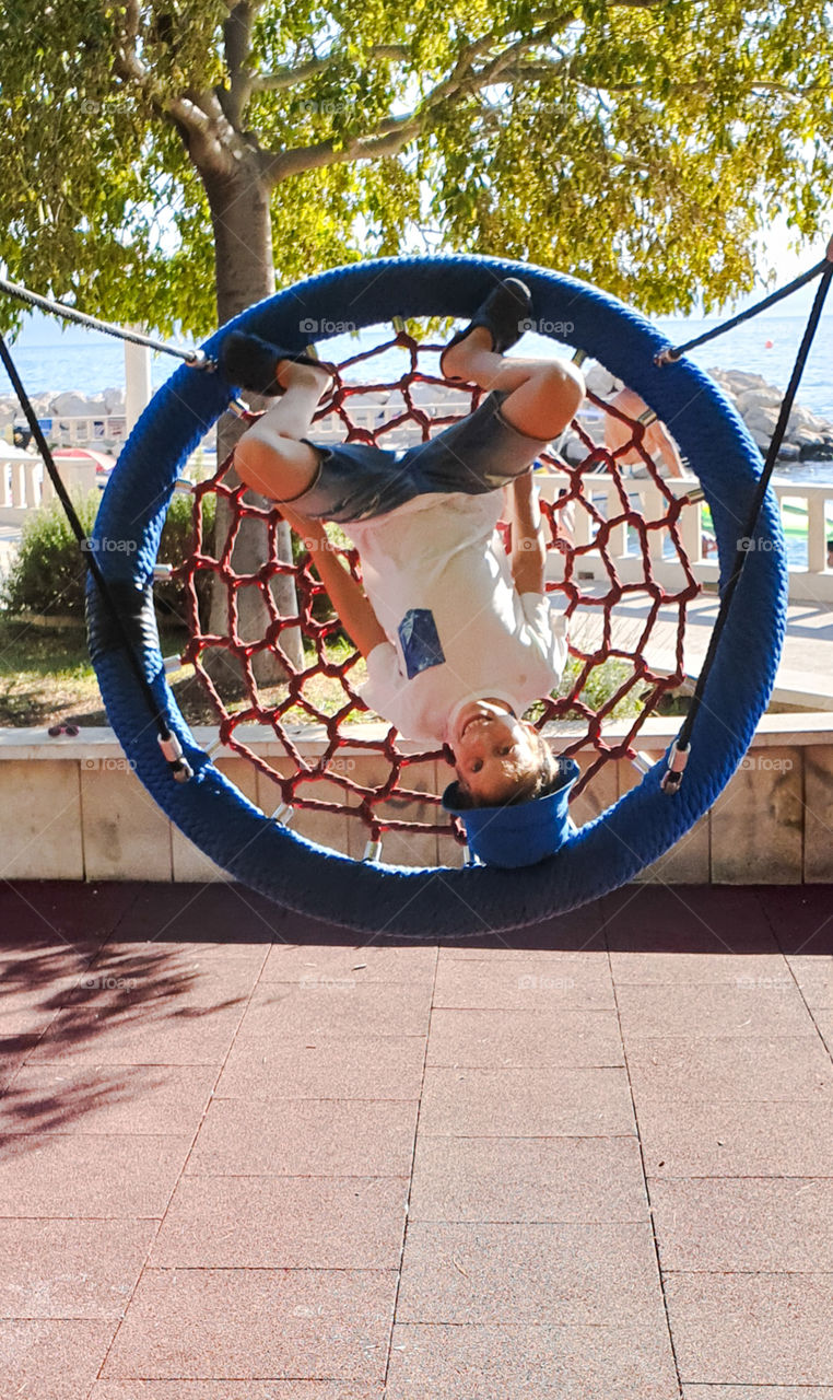 A boy riding a round swing upside down on a playground by the sea.  Summer Holidays, Croatia, Dalmatia, Tucepi