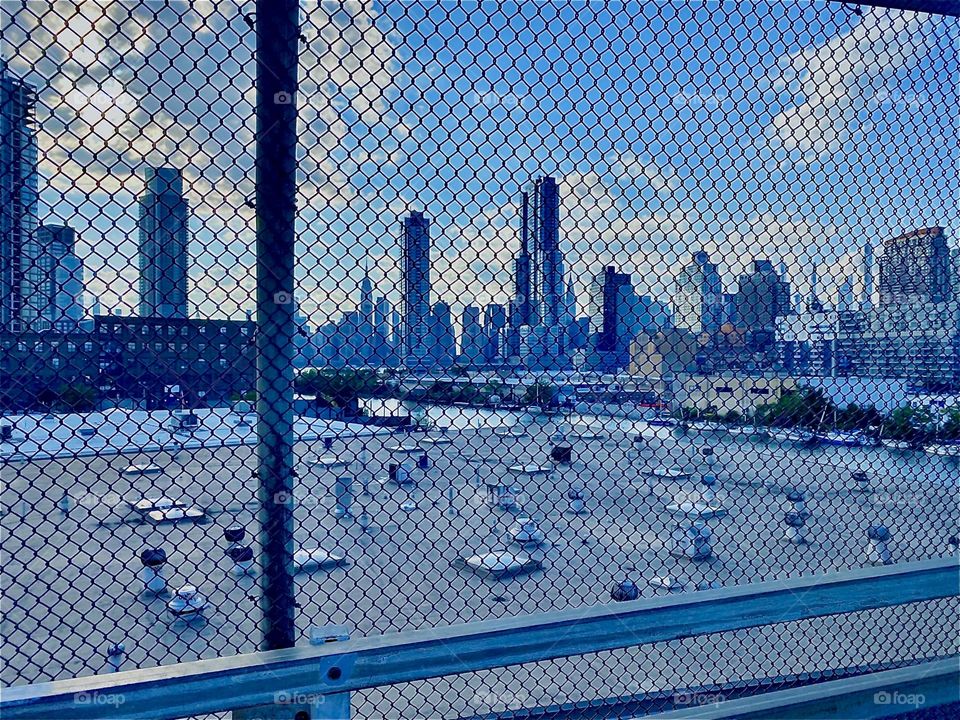 A large flat roof w skylights obstructs the view of the “E River” and we see the skylines of “LIC, Queens” & “Manhattan” thru a metal fence from the “Greenpoint, Bklyn” side of the bridge but it still is a nice photo. 2022. Hypnotic Productions