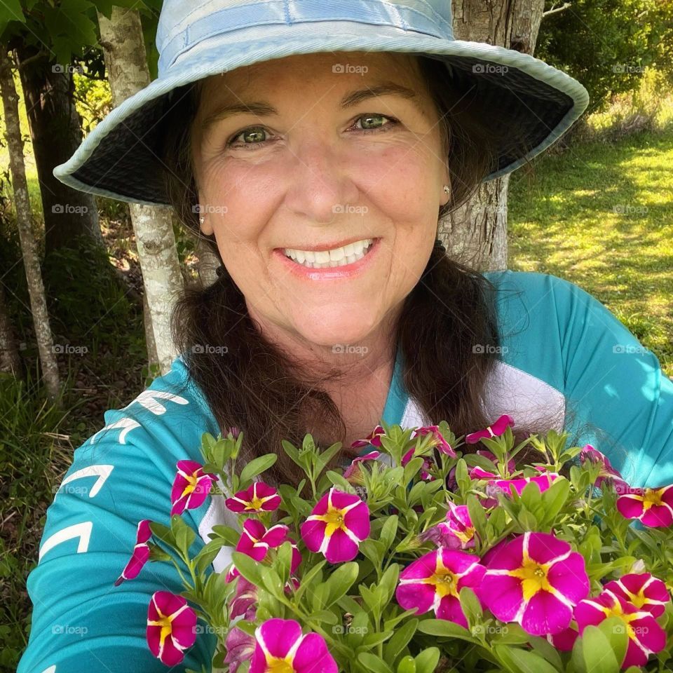 A smiling woman wearing a sun hat and holding an armful of colorful pink and yellow mini petunias ready to be planted in her Springtime garden.