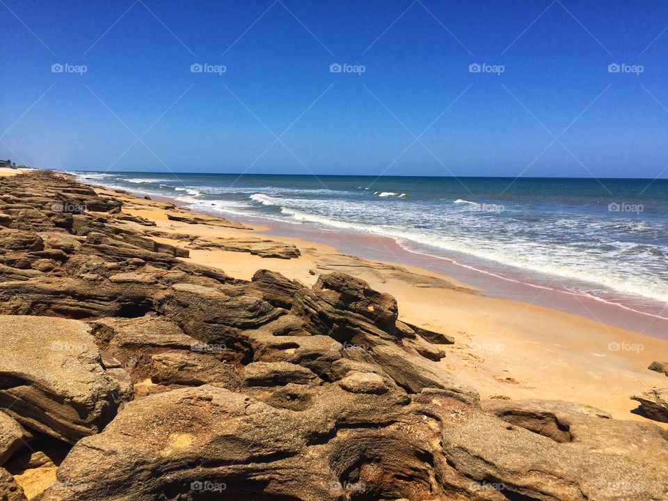 Waves crashing on a beach with rock formations under a bright blue cloudless sky