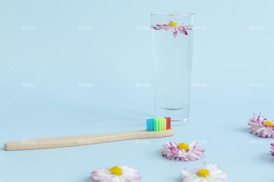 An ecological, wooden and reusable toothbrush with multi-colored bristles stands with a glass of water and a summer daisy, on a blue background.
