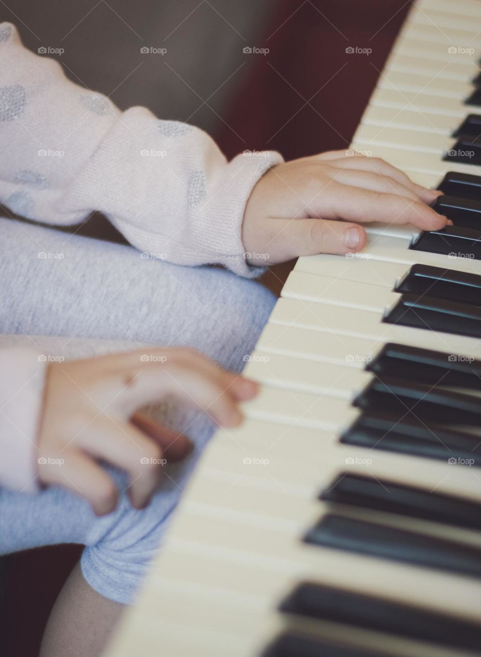 Hands of a little caucasian girl pressing her fingers on the keys of an electric piano while sitting on a sofa in the living room, close-up side view with selective focus. Music education concept, musical instruments.