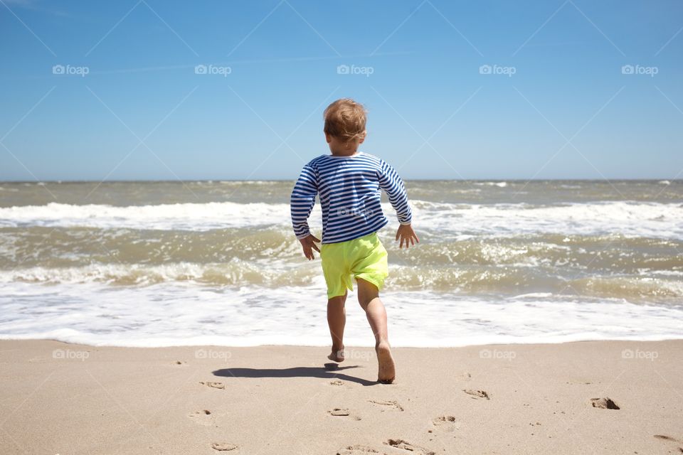 Little boy 3-4 years old in sailor striped shirt run to sea with waves and foam to swim, happy summer, vacation and childhood, back view 