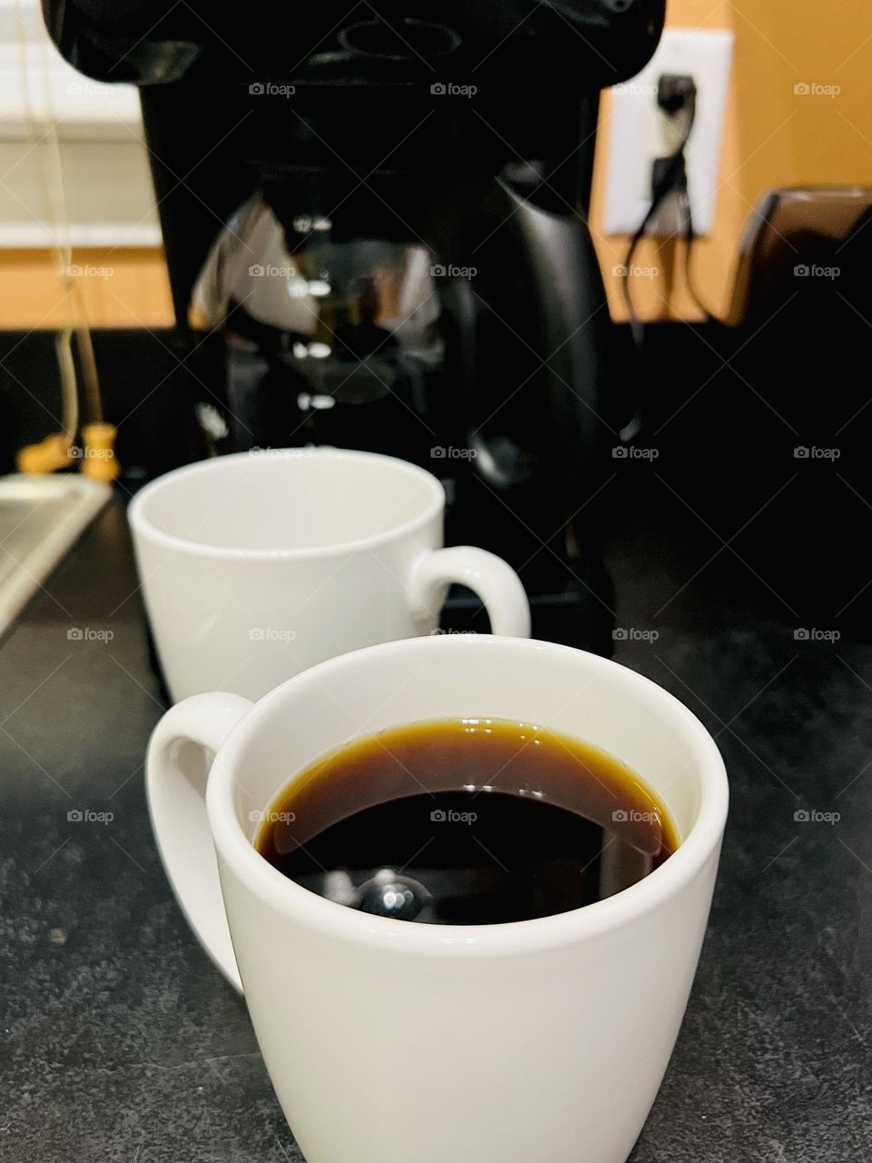 Overhead closeup of white coffee cup. Coffee is pouring from above into the half-filled mug in front of a black drip coffee maker. Setting is a home kitchen, a black laminate  counter with black appliances.