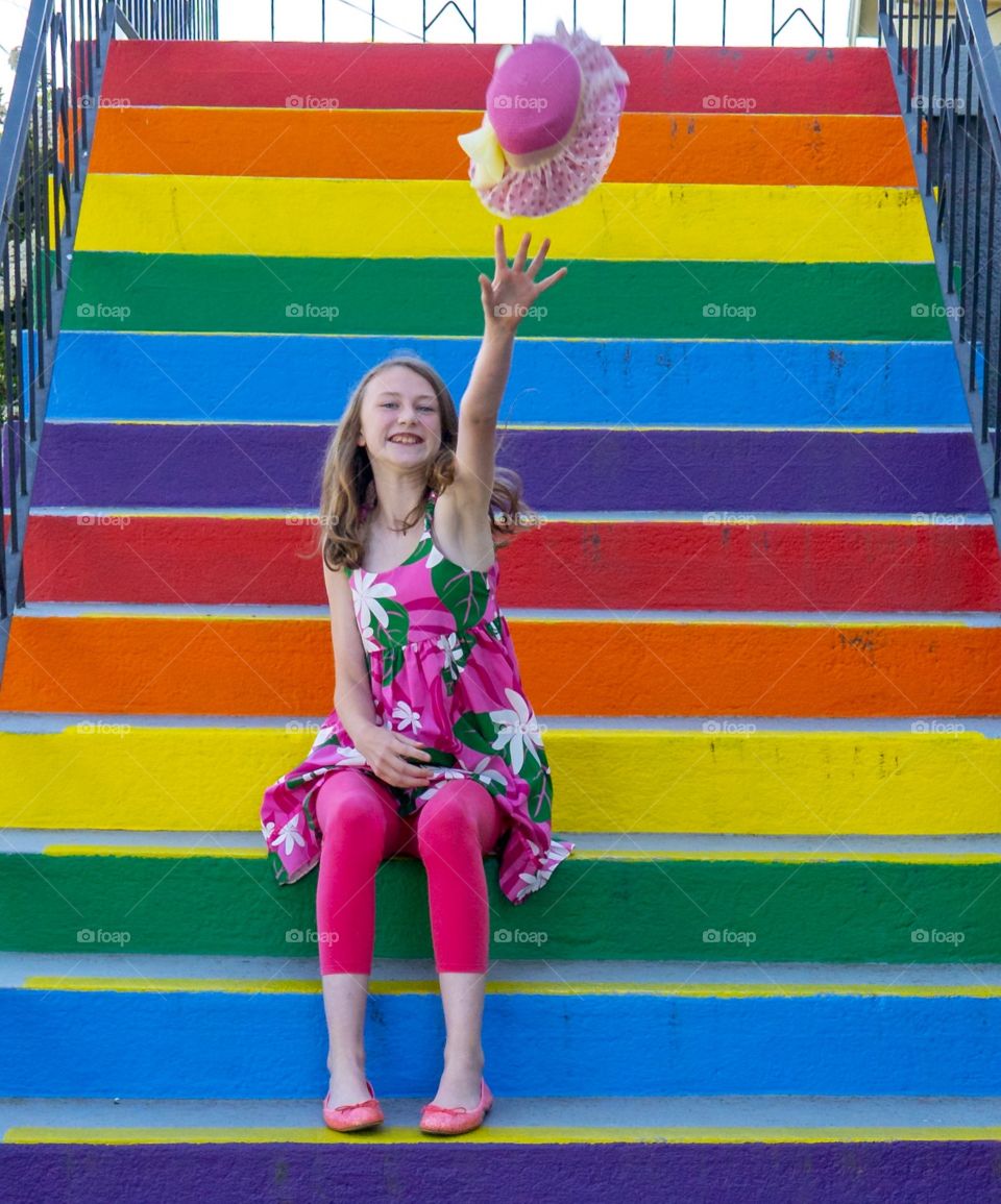 Girl tosses up hat while sitting on rainbow coloured stairs