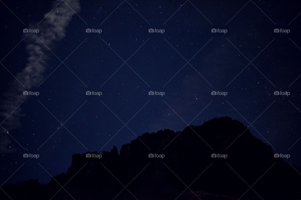 A beautiful night sky with clouds and many stars. A mountain creates layers and texture. Blue is the main color.