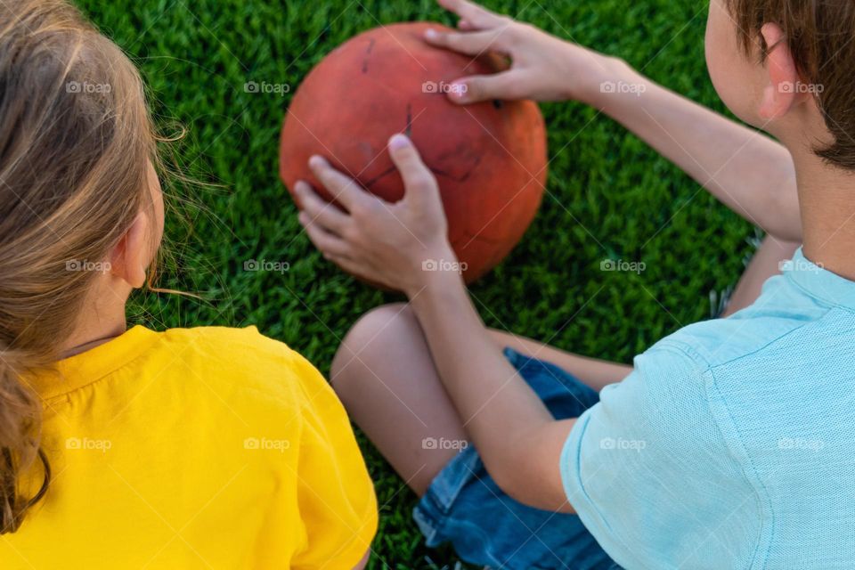 Back view of crop anonymous kids sitting on lawn and holding ball while spending time