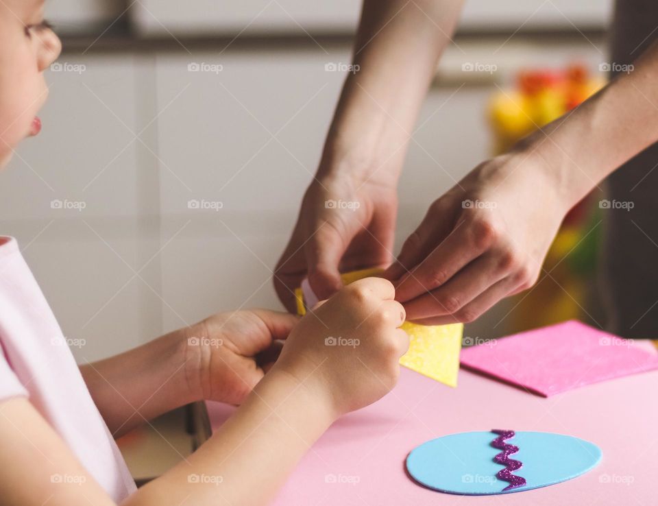 The hands of a young mother help a little beautiful Caucasian girl daughter peel off a felt sticker for crafts sitting at a children's table in the room, side view close-up. Creative kids concept.