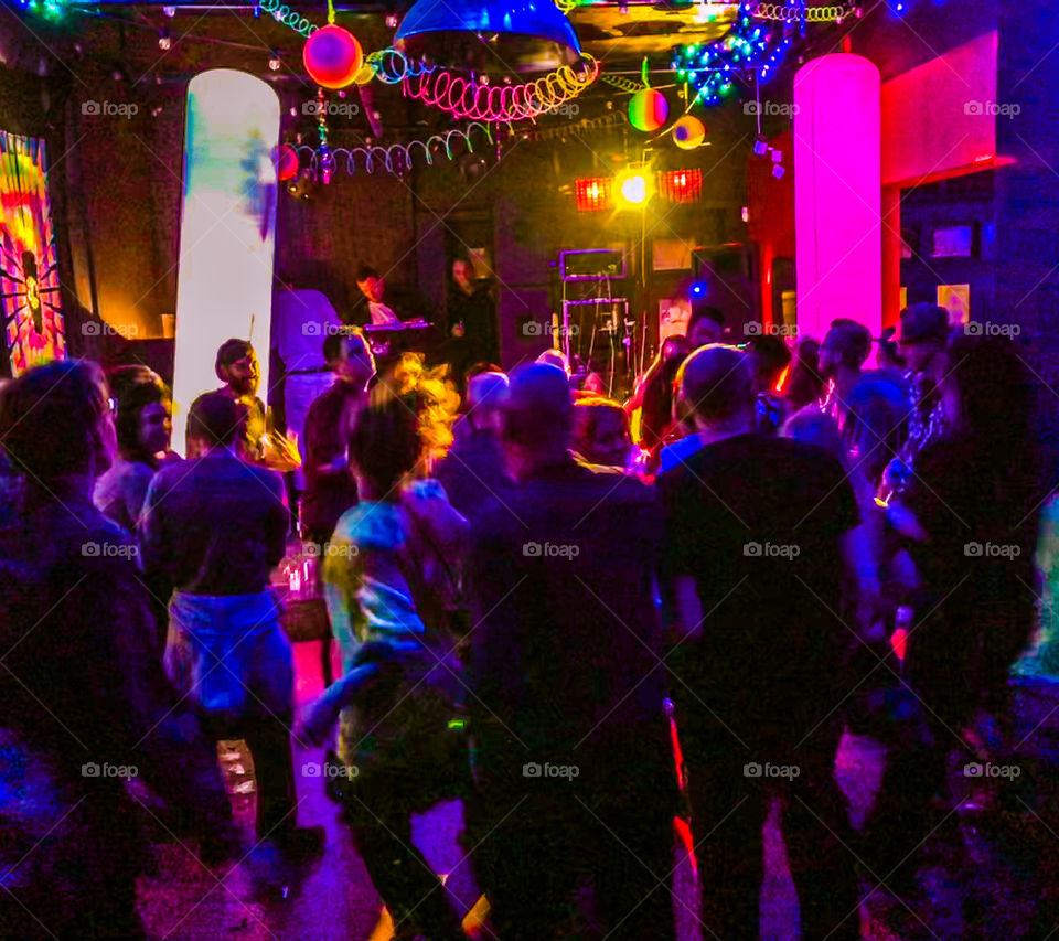 People dancing in a club, they are mostly silhouetted against the brightly coloured decor and lights, a DJ can be seen at the far end