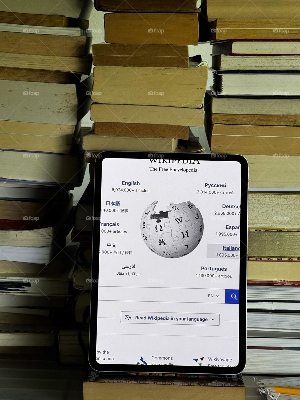 A stack of books with an iPad showing the Wikipedia start page next to a pencil and notepaper page