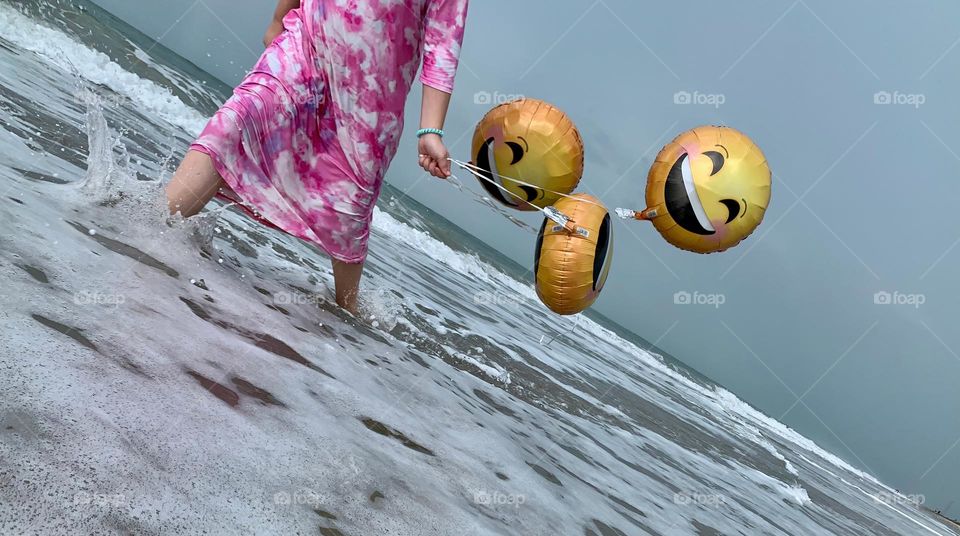 Little Girl Walking In The Water And Having Fun With Helium Emoji’s Balloons On The Beach By The Ocean.