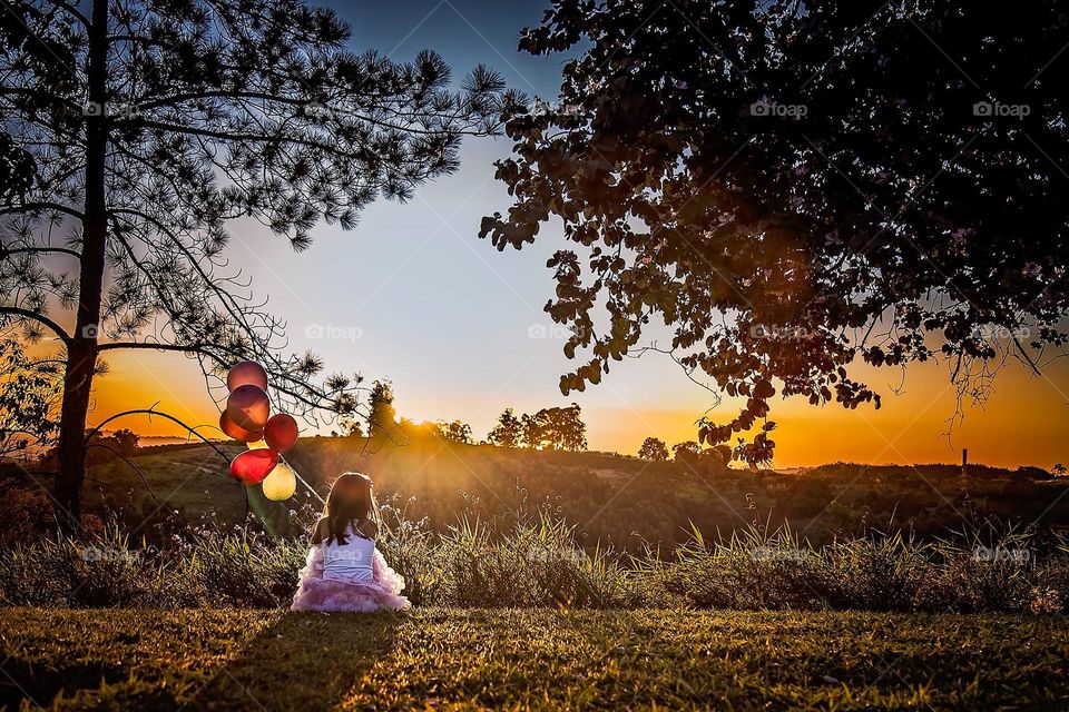 Harmonious composition in backlight: the child centers the scene while the trees frame the sunset, highlighting textures, golden light and balloons as a point of color