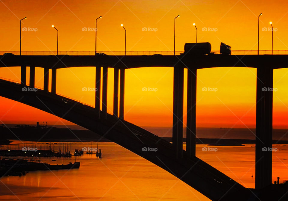 Traffic is silhouetted by the deep orange sunset over Ponte da Arrábida, a bridge that crosses Rio Douro in Porto - Portugal - Oct 2019