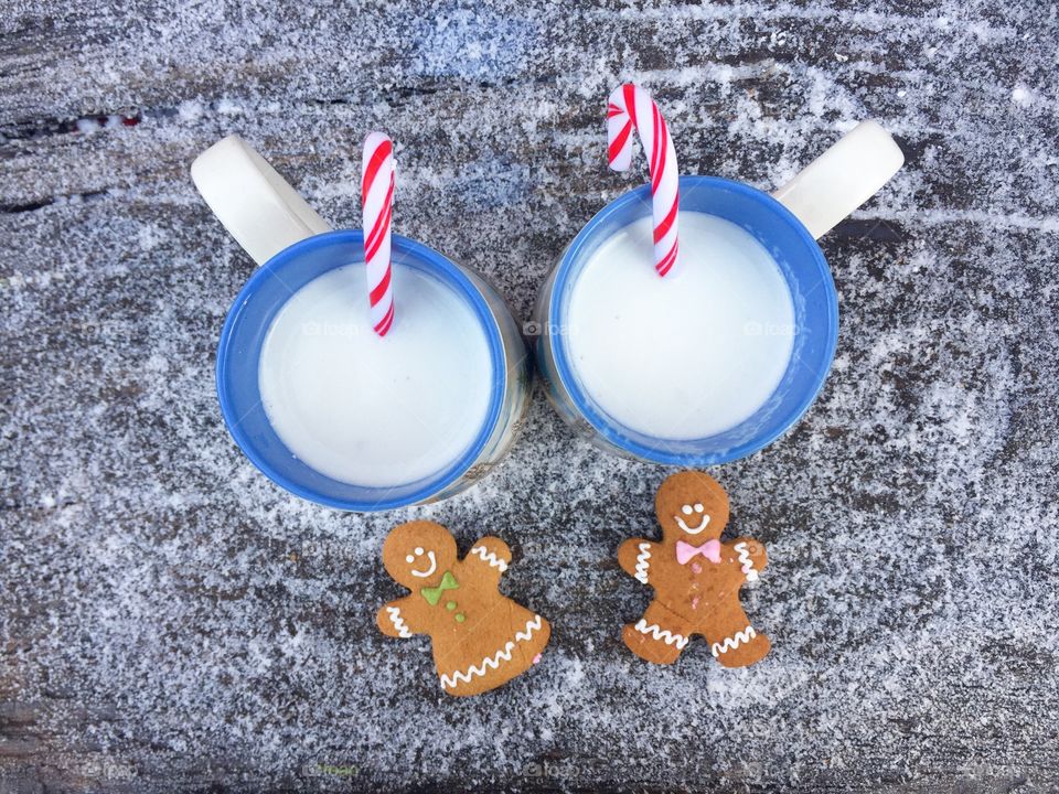 Two mugs of white chocolate and milk with candy canes inside on snowy table with two gingerbread men beside