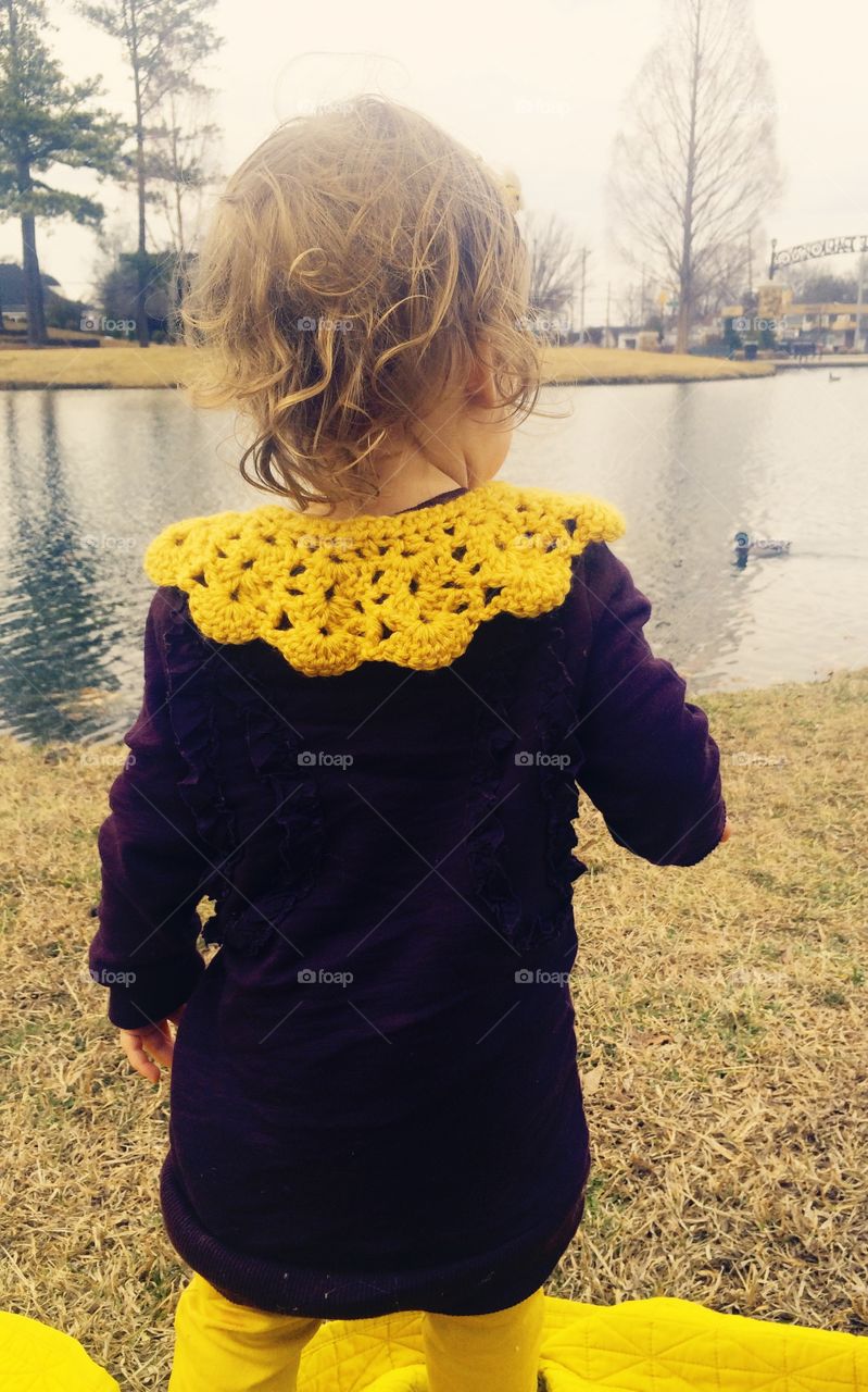 Blonde curly haired baby toddler looking at duck pond with rich, purple long sleeves and yellow crocheted collar 