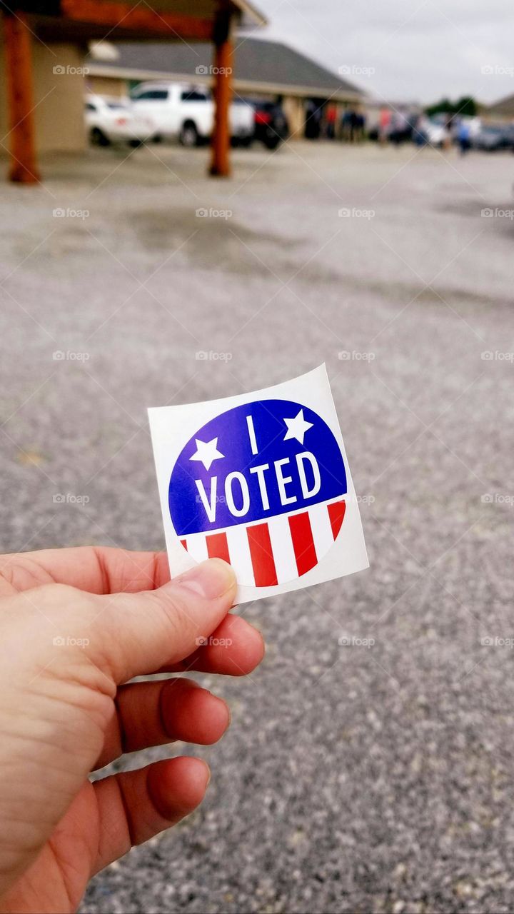 I Voted Stick in a Persons Hand standing in a parking lot with a line of people after voting in an American Election