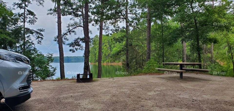 a view of the campsite that is lake side where we see the front of a silver vehicle, firepit, picnic table & some trees before the Lake & more trees in the distance