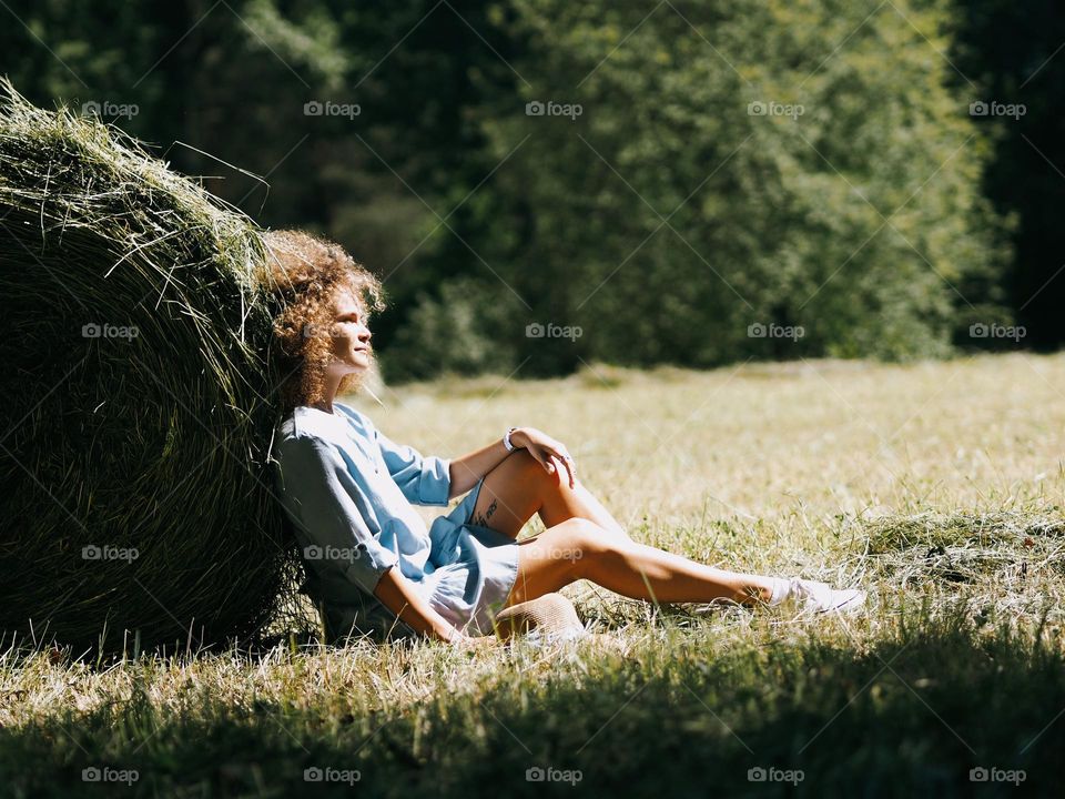 A young beautiful girl with curly hair sits on a green field near a stack of cut grass on a sunny summer day, glimmers 