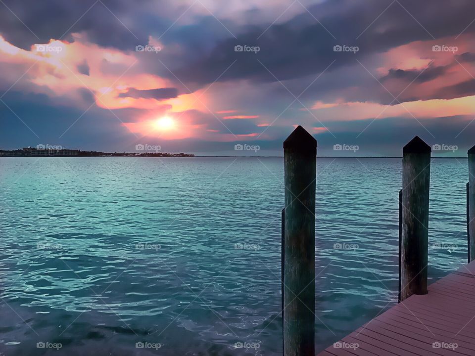 Purple clouds surround a red sunset as seen from a pier on the water.