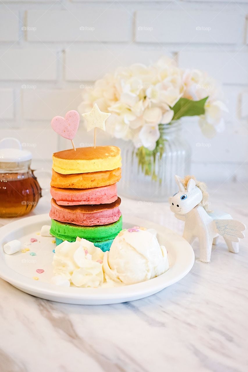 Rainbow pancakes served with vanilla ice cream and a unicorn wooden toy on the side of the dish. Maple syrup is on the background.