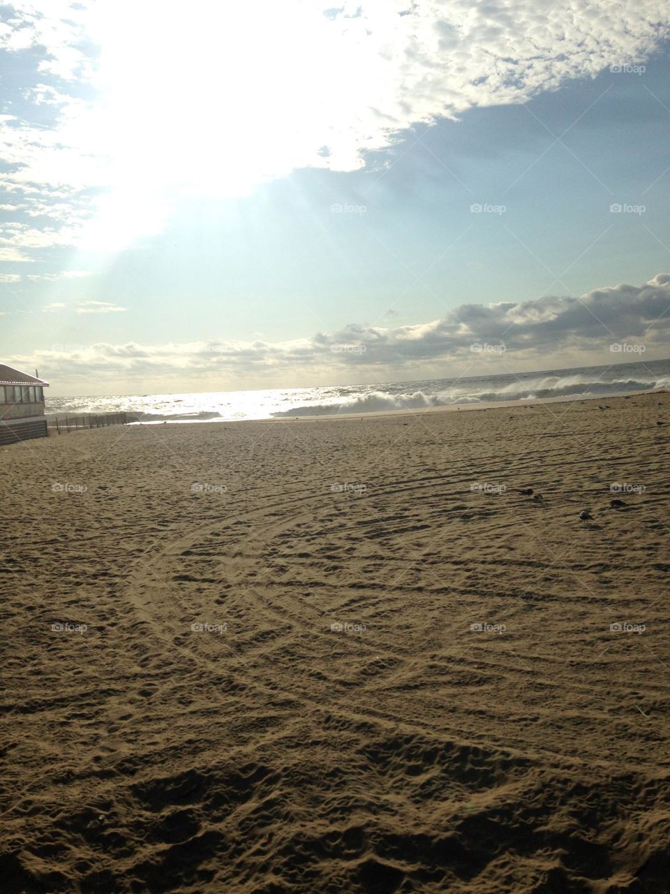 Tracks form a circular pattern in the sand in this photo of the beach taken from the boardwalk in Point Pleasant Beach, NJ. The wide stretch of sand is the focus of the photo, highlighted by ocean waves, clouds, and a streak of sun. 