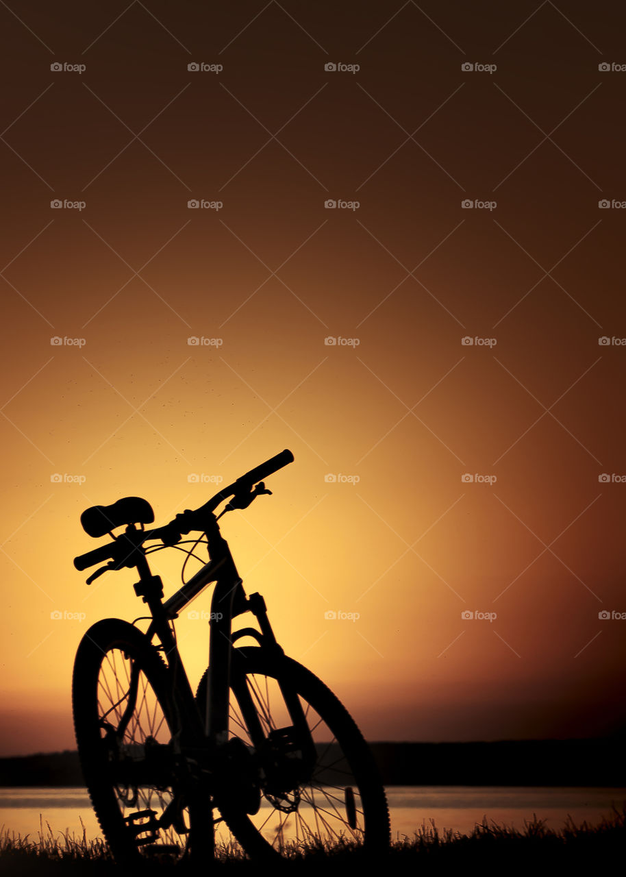 Dark silhouette of a single mountain bike, standing on a lakeshore during summer orange dusk