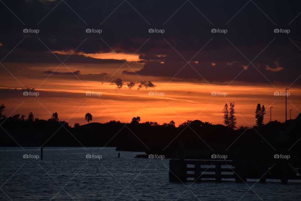 A view of the sunset looking out toward the water as the orange sky fades to black. The clouds have a purple tint. Silhouetted trees line the view in front of the sky. Taken near the beach in Florida.
