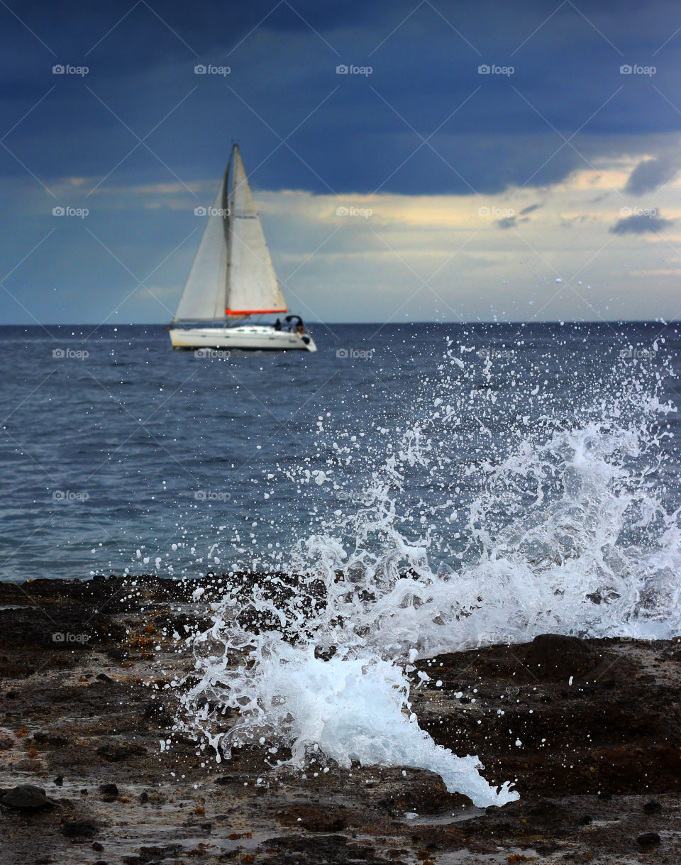 A Wave Crashes on a Rock in Front of a Sailboat