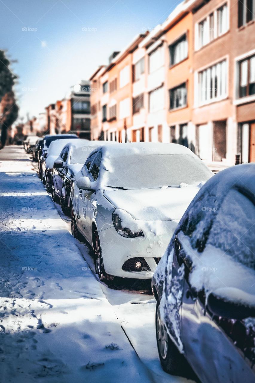 A row of snow-covered cars are parked on the roadside in the city of Brussels, Belgium, close-up top view.