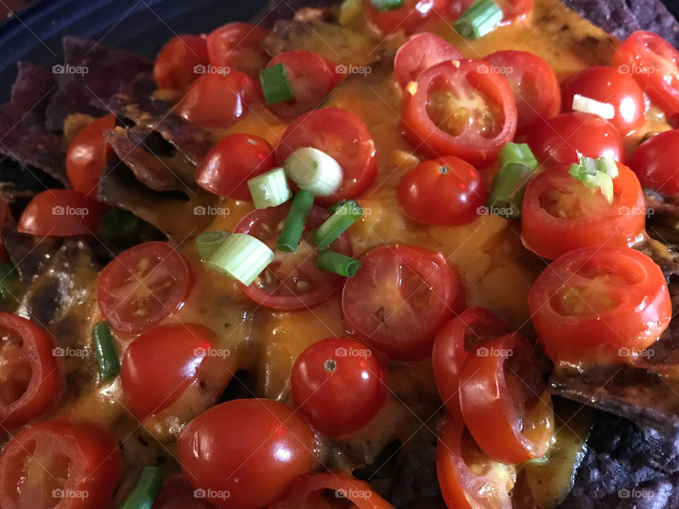 Who doesn’t like Nachos?! Closeup of blue corn chips, smothered with melted cheese, sliced grape tomatoes, green onions,  & a splash of hot sauce! Simple pero delicioso!