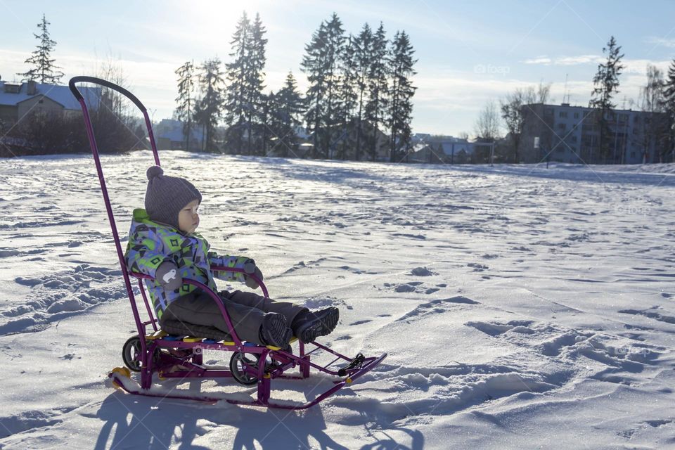 A child with a serious expression on his face in winter clothes jackets, pants, hat and boots in winter on white snow on the street and in the park in nature sledding and playing winter fun.