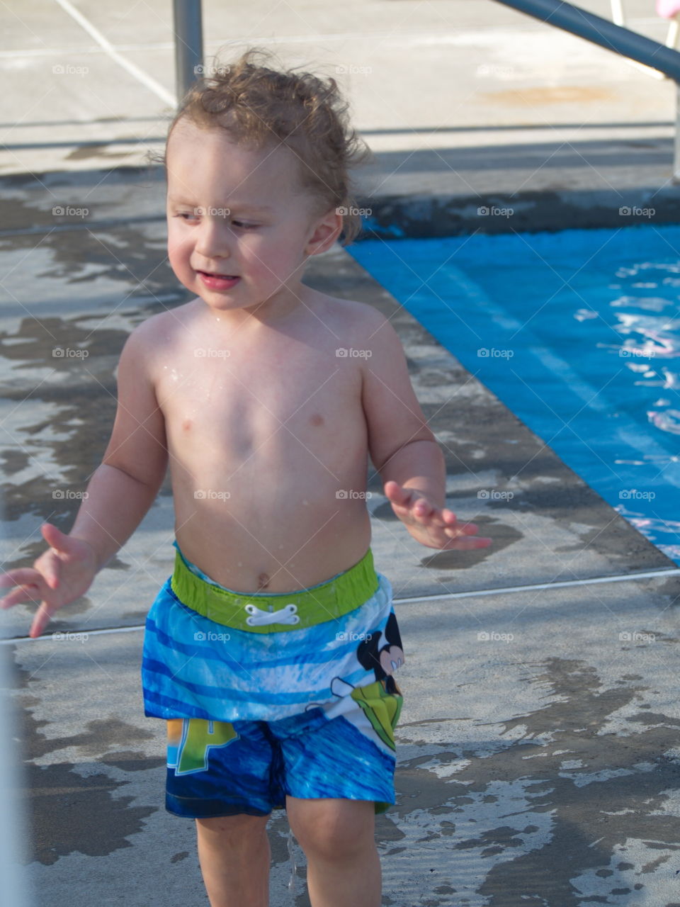 A cute little boy with an animated expression walks beside an outdoor pool on a sunny summer day. 