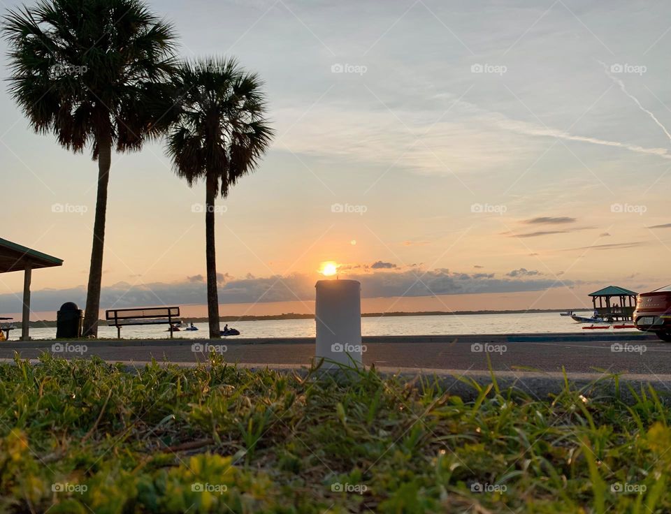 Between spring and summer time at sunset, the candle seems to lit up by the sun by the Indian lagoon river in Florida on the Central eastern side with tropical beautiful view with two palm trees and activity going on on the water.