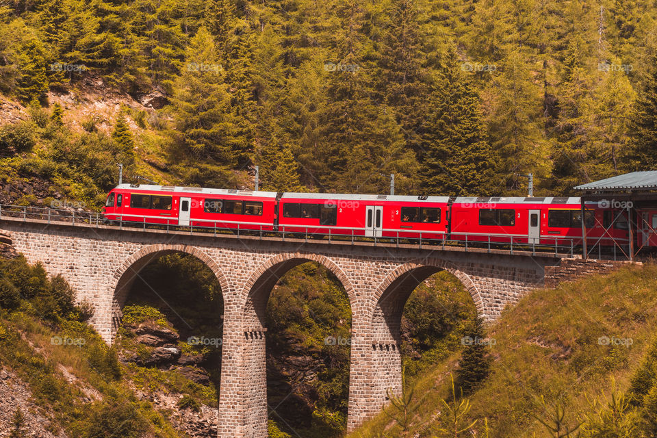 A red train crosses over an arch bridge in a forest mountain area in Switzerland 