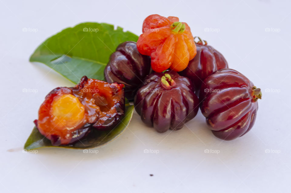 Cut open and whole single seed mature, ripe Pitanga cherries and leaf on white surface.