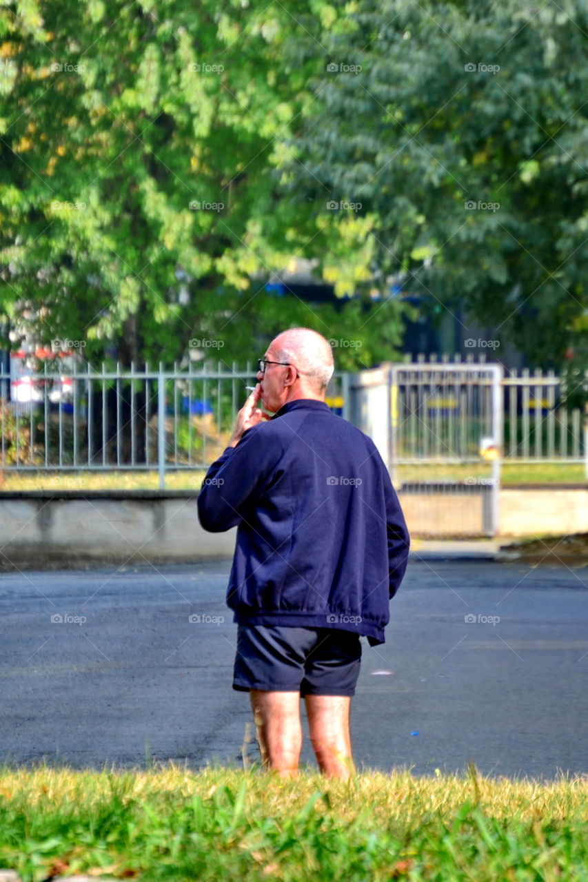 an elderly man in shorts, smoking a cigarette outdoors