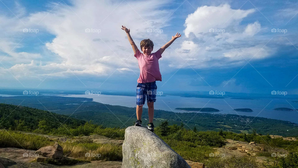 Young boy standing on a Boulder atop a mountain with his arms up. Distant lake scene behind him.