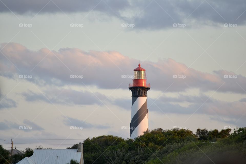 A black, white, and red striped lighthouse towering above green trees and a body of water against a blue purple and pink dusk sky