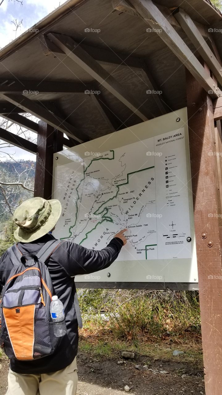 man wearing a backpack and a hat pointing at a trail map for a mountain hiking during the winter, spring, summer, fall season